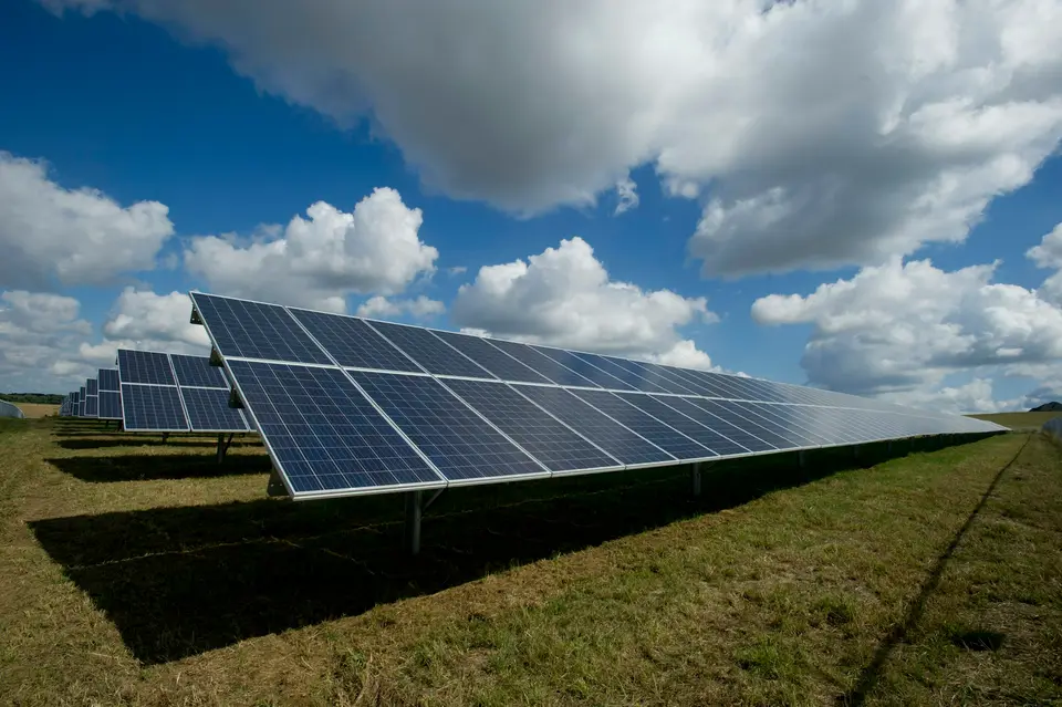 Rows of solar panels in a green field under a bright, cloud-filled sky